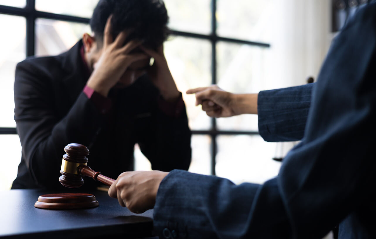 A person sits with their head in their hands, while another person points and holds a gavel, symbolizing a legal confrontation.